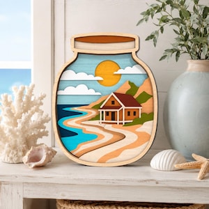 May include: Wooden layered art piece depicting a beach scene inside a jar shape. The artwork features a house, sun, clouds, and ocean waves in shades of orange, blue, and beige. The piece is displayed on a white surface.