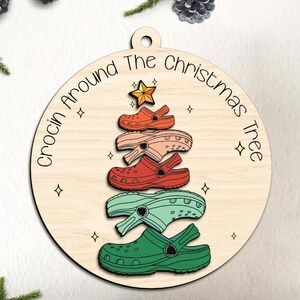Puede incluir: Un adorno navideño de madera con forma de árbol de Navidad hecho de Crocs coloridos apilados. El adorno está decorado con estrellas y el texto "Crocin Around The Christmas Tree".