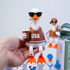 May include: A white duck figurine wearing black sunglasses and a brown jersey with the text USA printed in white. The duck holds a brown coffee cup and has articulated orange webbed feet. In the background, another duck wears a brown cap and holds a flag.