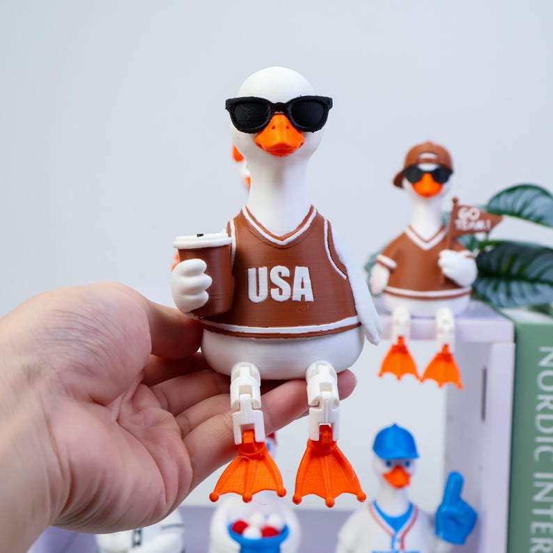 May include: A white duck figurine wearing black sunglasses and a brown jersey with the text USA printed in white. The duck holds a brown coffee cup and has articulated orange webbed feet. In the background, another duck wears a brown cap and holds a flag.
