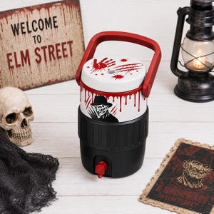 May include: A black and white beverage cooler with a red handle and spigot. The white upper section features a graphic of a character with a fedora and clawed hand, accented by red blood-like drips, a handprint, and splatter patterns.