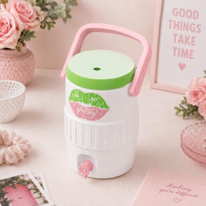 Op de afbeelding: Een witte drankkoeler met een lichtgroen deksel en een roze handvat. De koeler is voorzien van een afbeelding van lippen in groen en roze met de tekst "oh so pretty" in het midden. Een kleine roze kraan bevindt zich aan de onderkant.
