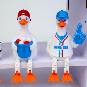 May include: Two white duck figurines with orange webbed feet. The left duck wears a red cap and holds a blue bucket filled with white and red balls. The right duck wears a blue cap and a white baseball jersey with the text HOME RUN, holding a blue foam finger.