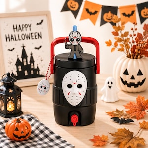 May include: A black beverage cooler featuring a white hockey mask on the front, a red handle, and a small Jason Voorhees figure on the lid. A matching hockey mask charm hangs from a red beaded chain on the side of the cooler, which has a red spigot.
