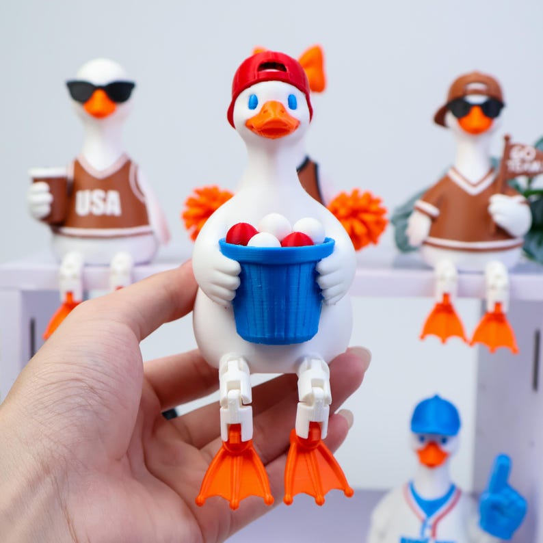 May include: A white duck figurine with an orange beak and feet, wearing a red baseball cap, holding a blue bucket filled with small red and white balls. The duck has articulated legs and is part of a collection of similar duck figures.
