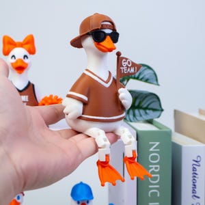 May include: A white duck figurine wearing a brown baseball cap, black sunglasses, and a brown jersey with white trim. The duck holds a small brown flag with the text GO TEAM! in white. It has articulated white legs and bright orange webbed feet.