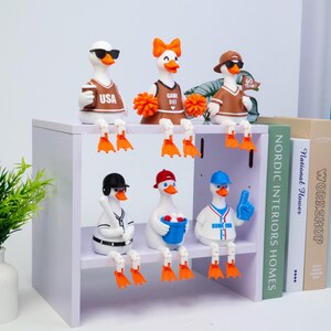 May include: Six white duck figurines with orange beaks and feet, arranged on two shelves. Top row: a duck in sunglasses with a USA jersey, a cheerleader duck with orange pom-poms, and a duck in a cap with a Go Team flag. Bottom row: a baseball duck, a duck with a bucket of balls, and a duck in a baseball jersey with a blue foam finger.
