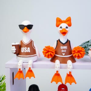 May include: Two white duck figurines with orange beaks and feet. The left duck wears black sunglasses and a brown jersey with USA printed on it, holding a brown cup. The right duck wears an orange bow and a brown jersey with GAME DAY printed on it.