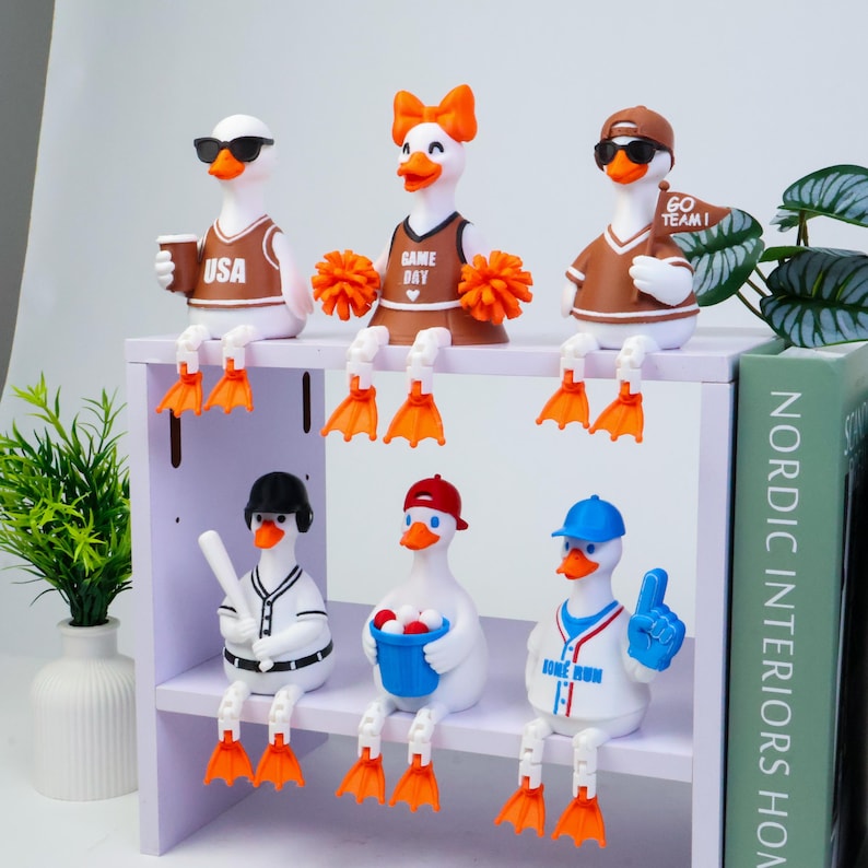 May include: Six white duck figurines with orange beaks and feet, posed on a shelf. Top row: a duck in a USA jersey with sunglasses, a cheerleader duck with orange pom-poms, and a duck in a brown jersey holding a GO TEAM flag. Bottom row: a baseball player duck, a duck holding a bucket of balls, and a duck in a baseball jersey with a foam finger.
