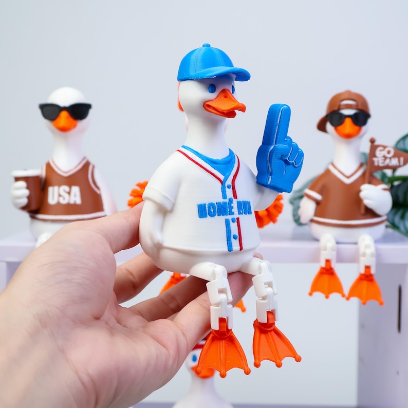 May include: A white rubber duck figurine wearing a blue baseball cap and a white jersey with the text HOME RUN in blue. The duck has orange feet and holds a blue foam finger. Two other duck figurines are visible in the background, one with USA text.