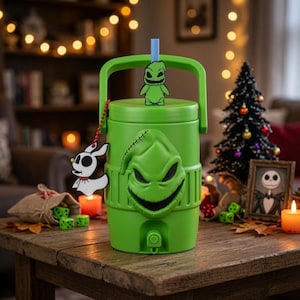 May include: A bright green cylindrical beverage cooler featuring a moulded relief of a grinning character face on the front. It has a top handle, a light blue straw, and a small white ghost-shaped charm attached by a red beaded chain.