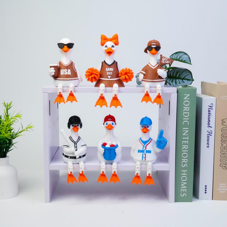 May include: Six white duck figurines with orange beaks and feet, arranged on a two-tier shelf. The top row features ducks in brown and white sports attire, including a cheerleader with orange pom-poms. The bottom row shows ducks in baseball gear.