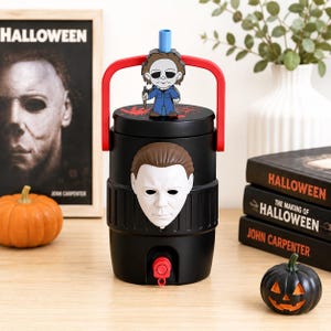 May include: A black cylindrical beverage cooler featuring a white Michael Myers mask on the front, a red handle, and a red spigot at the base. The lid displays a red handprint and a small Michael Myers figure attached to a blue straw.