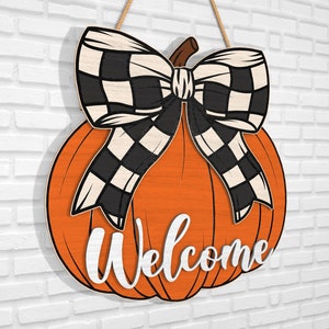 Puede incluir: Un colgador de puerta de madera en forma de calabaza con un lazo a cuadros blanco y negro y la palabra "Bienvenido" escrita en la parte delantera.