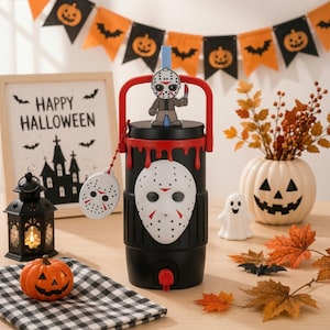 May include: A black beverage dispenser featuring a white hockey mask emblem on the front, a red handle, and a red spigot. A small hockey mask charm hangs from a red chain, and a cartoon figure of a masked character sits on the lid straw.
