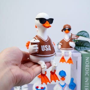 May include: A white duck figurine wearing black sunglasses and a brown jersey with the text USA printed in white. The duck holds a brown cup in one wing and has bright orange feet. Other similar duck figurines are visible in the background.