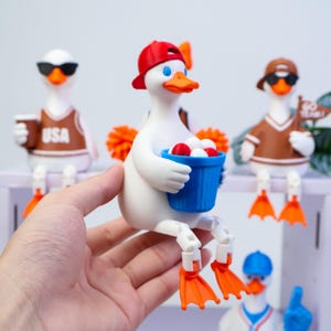 May include: A white duck figurine wearing a red baseball cap and holding a blue bucket filled with small white and red balls. The duck has orange feet and a beak, with articulated legs that allow it to sit on an edge. Other duck figurines are visible.