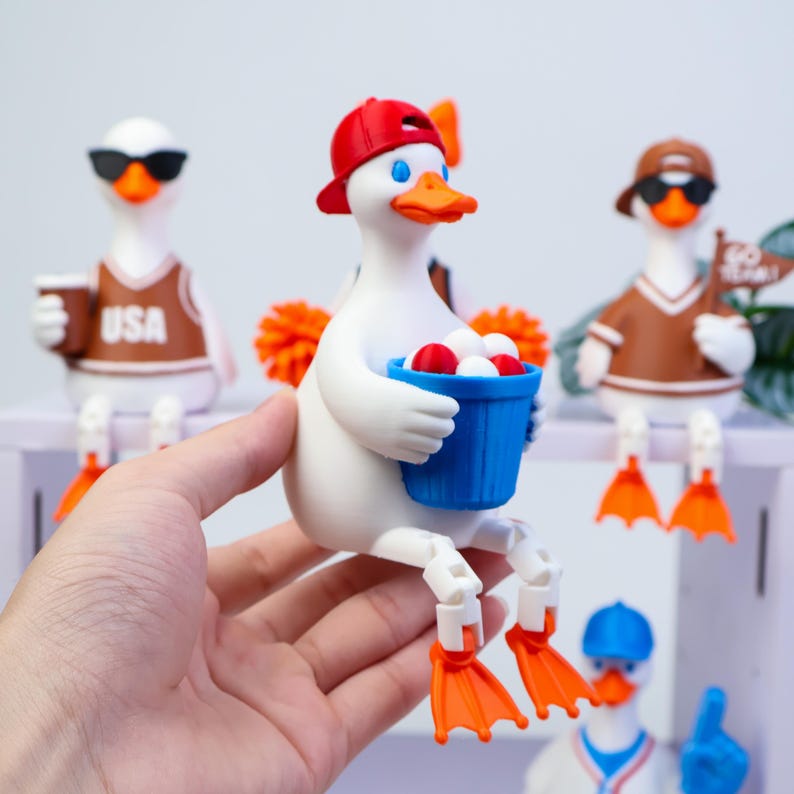 May include: A white duck figurine wearing a red baseball cap and holding a blue bucket filled with small white and red balls. The duck has orange feet and a beak, with articulated legs that allow it to sit on an edge. Other duck figurines are visible.