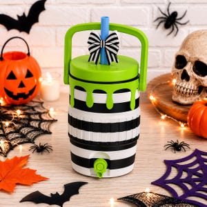 May include: A cylindrical beverage dispenser featuring a black and white horizontal striped pattern with a bright green lid and handle. The lid has a green drip design and a blue straw topped with a black and white striped bow. A green spigot is at the base.