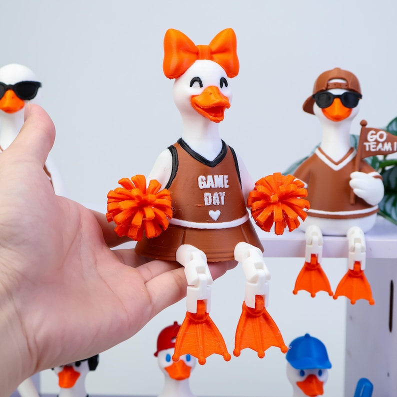 May include: A white duck figurine wearing an orange bow, a brown cheerleading uniform with the text GAME DAY and a heart, and orange pom-poms. The duck has articulated white legs with orange webbed feet. Other duck figurines are visible nearby.