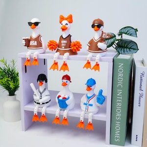 May include: Six white duck figurines with orange beaks and feet, dressed in sports-themed outfits. The top row features ducks with sunglasses, a cheerleader with pom-poms, and a fan with a flag. The bottom row includes baseball-themed ducks.