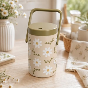 May include: A cylindrical beige container with a solid olive green lid and a matching arched handle. The exterior features a repeating pattern of white daisy flowers with yellow centers and small olive green leafy vine accents.