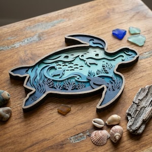 Op de afbeelding: Een meerlaags houten kunstwerk in de vorm van een zeeschildpad, met een gedetailleerde onderwaterwereld. De lagen hout tonen tinten blauw en groen, die golven, vissen en koraal uitbeelden. Schelpen en zeeglas sieren het oppervlak.