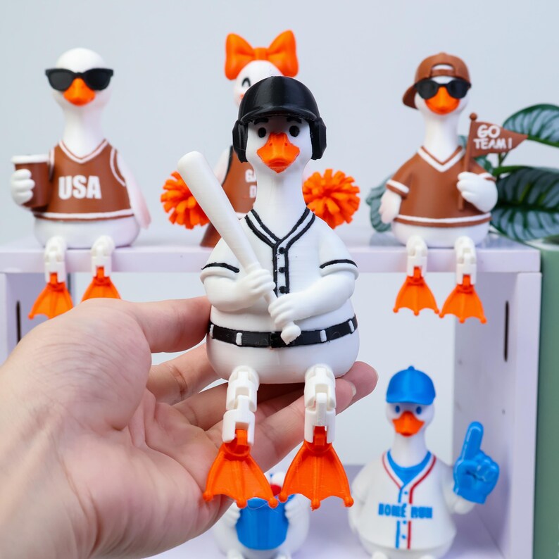 May include: A white duck figurine wearing a black baseball cap and a white baseball uniform with black trim, holding a white baseball bat. The duck has orange feet and is part of a collection of similar duck figurines with various sports themes.