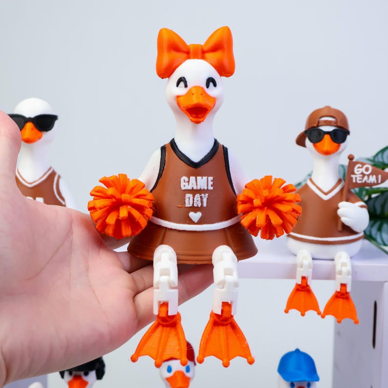 May include: A white duck figurine dressed as a cheerleader in a brown uniform with the text GAME DAY and a heart icon. The duck has an orange bow, orange pom-poms, and orange webbed feet. Two other duck figurines are visible in the background.