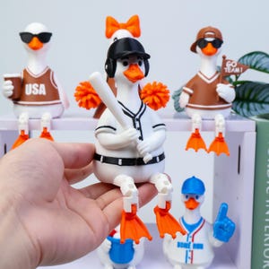May include: A white duck figurine wearing a black baseball helmet and a white baseball uniform with black trim, holding a white baseball bat. The duck has orange feet and is positioned in a seated pose with articulated legs hanging down.