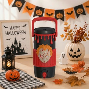 May include: A black and red beverage dispenser featuring a sculpted, scarred doll face on the front. The top section has a red dripping design, a black lid, and a red carrying handle. A red spigot is located at the bottom of the cylindrical container.