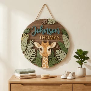 Könnte beinhalten: Ein rundes Holzschild mit einem Giraffen-Design und dem Namen "Johnson Thomas" in blauen und goldenen Buchstaben. Das Schild ist von grünen und beigen Blättern umgeben und hat eine Schnur zum Aufhängen. Ein Stapel gefalteter Tücher, eine Bürste und Babyschuhe liegen auf einer Holzoberfläche.