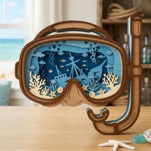 May include: A wooden decorative piece shaped like a diving mask and snorkel. The mask's interior features a layered design depicting an underwater scene with a sunken ship, fish, and coral in shades of blue and beige.