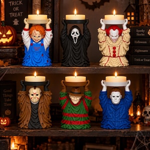 May include: Six horror movie character candle holders, each holding a lit pillar candle above their head. The figures include Chucky, Ghostface, Pennywise, Jason Voorhees, Freddy Krueger, and Michael Myers, all rendered in detailed, colorful resin.