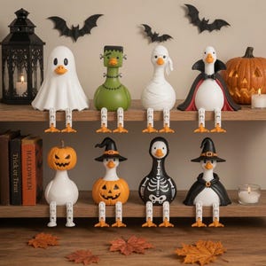 May include: A collection of eight duck figurines dressed in Halloween costumes, including a ghost, Frankenstein's monster, a mummy, a vampire, a pumpkin-headed duck, a pumpkin-bodied duck, a skeleton, and a witch, all with dangling legs.