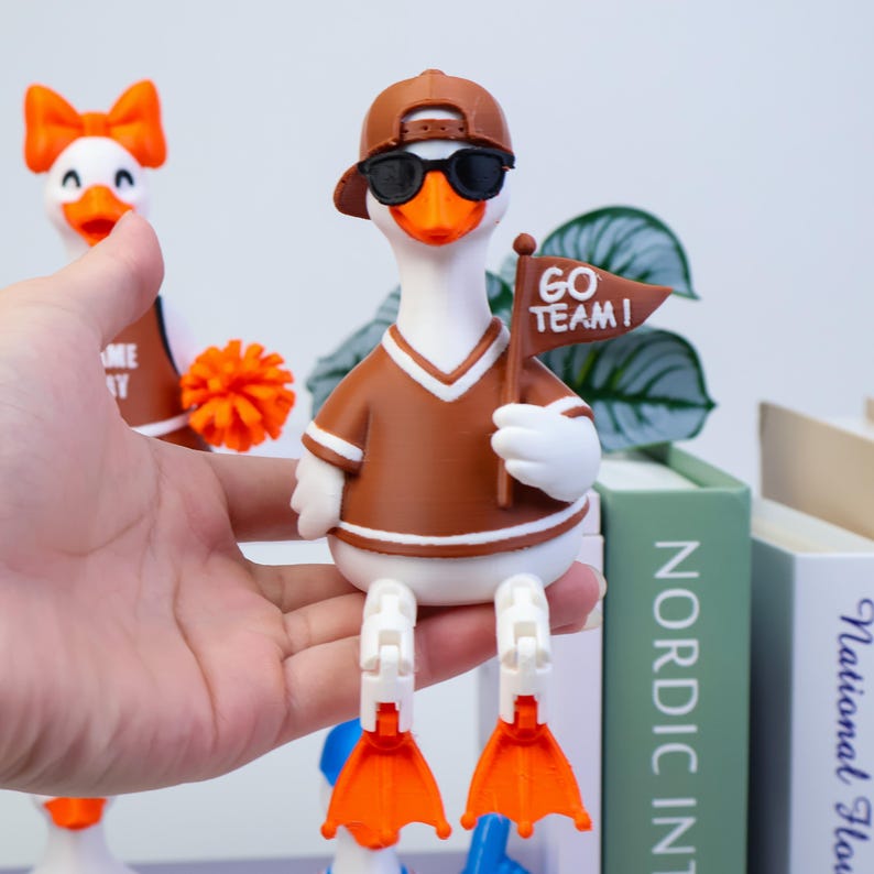 May include: A white duck figurine wearing a brown baseball cap, black sunglasses, and a brown jersey with white trim. The duck holds a brown pennant flag with the white text GO TEAM! and has articulated white legs with bright orange webbed feet.