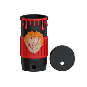 May include: A black cylindrical tumbler featuring a raised, three-dimensional orange-haired doll face with facial scars. The top rim has red dripping paint effects, and a red textured band wraps around the center. A separate black circular lid is included.
