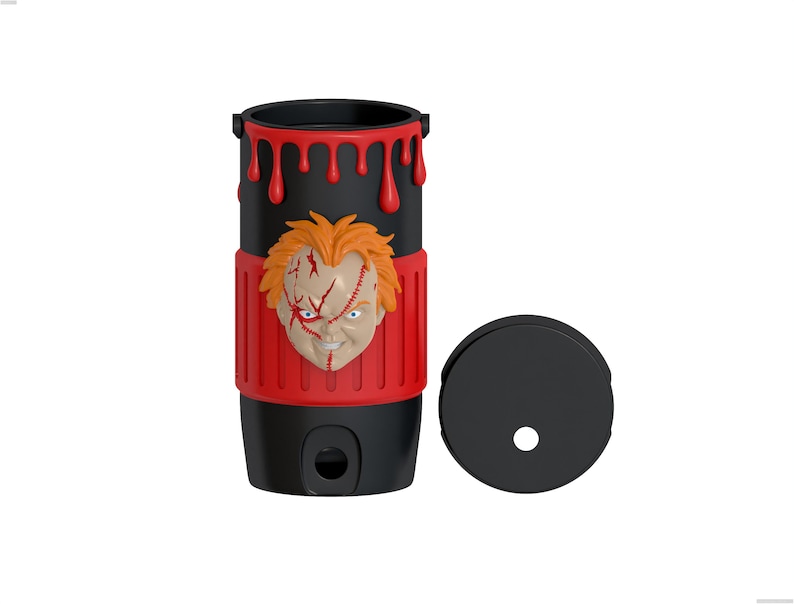 May include: A black cylindrical tumbler featuring a raised, three-dimensional orange-haired doll face with facial scars. The top rim has red dripping paint effects, and a red textured band wraps around the center. A separate black circular lid is included.