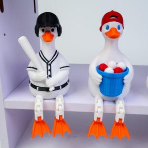 May include: Two white duck figurines with orange feet and articulated legs. The left duck wears a black baseball helmet and holds a white bat. The right duck wears a red baseball cap and holds a blue bucket filled with white and red balls.