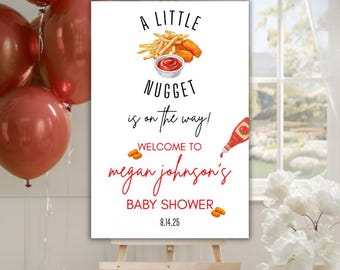 May include: A white sign with black text announcing a baby shower for Megan Johnson. The sign features a cartoon image of chicken nuggets and french fries with a red dipping sauce. The text reads "A Little Nugget is on the way! Welcome to Megan Johnson's Baby Shower 8.14.25".