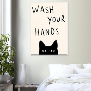 Pode incluir: Uma impressão de arte de parede retangular com fundo creme e o texto manuscrito em preto WASH YOUR HANDS. Abaixo do texto está a silhueta preta de uma cabeça de gato com dois olhos brancos espreitando sobre uma linha horizontal fina.