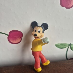 K&ouml;nnte beinhalten: Eine Mickey Mouse-Figur in Tanzpose. Die Figur hat schwarze Ohren, ein gelbes Hemd, eine rote Hose und gelbe Schuhe. Der Hintergrund zeigt eine Tapete mit Kirschmuster.