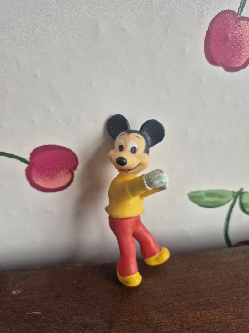 K&ouml;nnte beinhalten: Eine Mickey Mouse-Figur in Tanzpose. Die Figur hat schwarze Ohren, ein gelbes Hemd, eine rote Hose und gelbe Schuhe. Der Hintergrund zeigt eine Tapete mit Kirschmuster.
