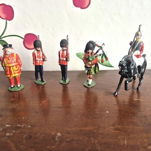 May include: A collection of vintage toy soldiers. The figures include a Beefeater in a red and gold uniform, soldiers in red jackets and black trousers, a bagpiper in a kilt, and a mounted soldier on a black horse.