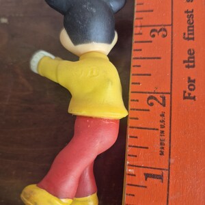 K&ouml;nnte beinhalten: Eine Vintage-Mickey-Mouse-Figur mit schwarzen Ohren, einer gelben Jacke, einer roten Hose und gelben Schuhen. Die Figur ist etwa 5 cm gro&szlig;. Ein Holzlineal befindet sich neben der Figur.