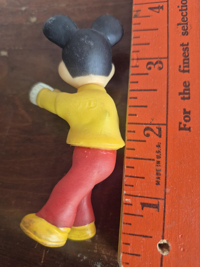 K&ouml;nnte beinhalten: Eine Vintage-Mickey-Mouse-Figur mit schwarzen Ohren, einer gelben Jacke, einer roten Hose und gelben Schuhen. Die Figur ist etwa 5 cm gro&szlig;. Ein Holzlineal befindet sich neben der Figur.
