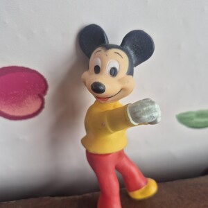 K&ouml;nnte beinhalten: Eine Vintage-Mickey-Mouse-Figur. Die Figur tr&auml;gt ein gelbes Hemd, eine rote Hose und gelbe Schuhe. Mickey hat schwarze Ohren und ein l&auml;chelndes Gesicht. Die Figur ist mit einem ausgestreckten Arm posiert.