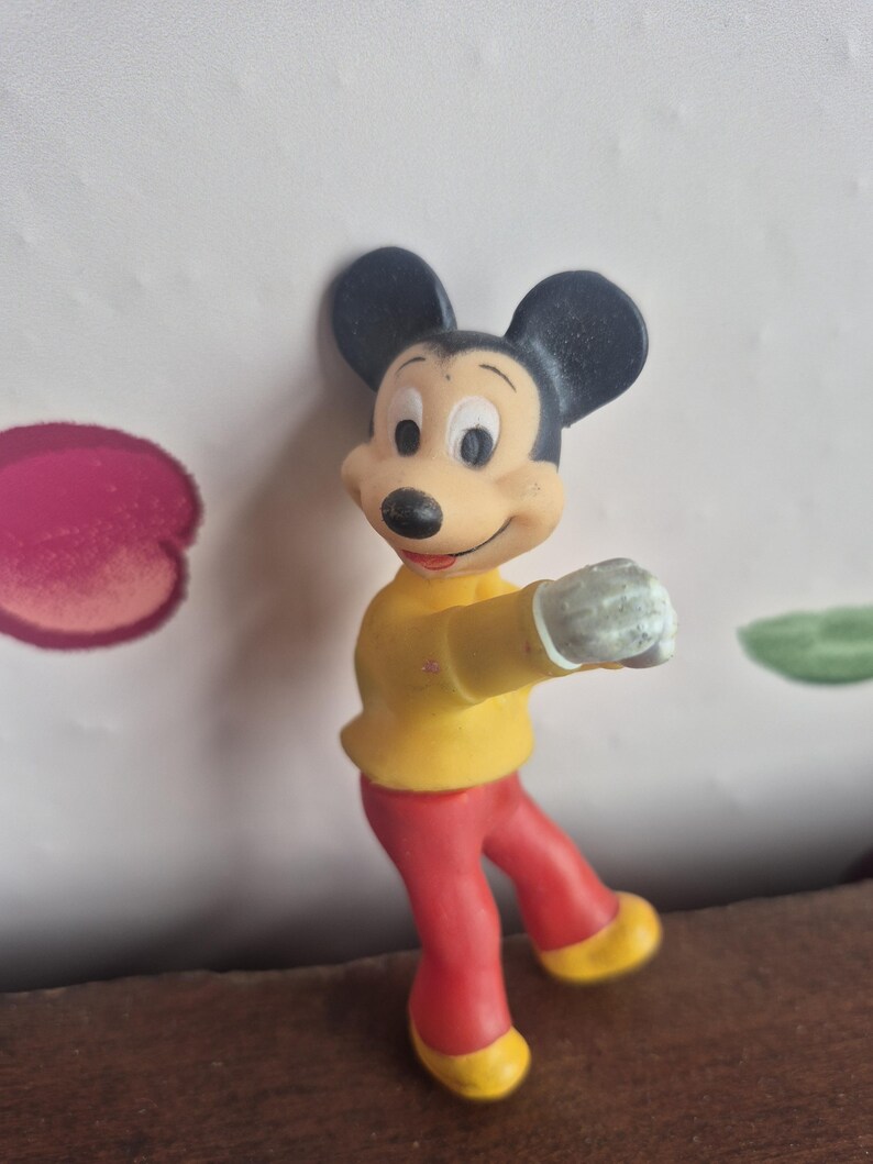K&ouml;nnte beinhalten: Eine Vintage-Mickey-Mouse-Figur. Die Figur tr&auml;gt ein gelbes Hemd, eine rote Hose und gelbe Schuhe. Mickey hat schwarze Ohren und ein l&auml;chelndes Gesicht. Die Figur ist mit einem ausgestreckten Arm posiert.
