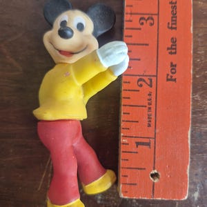K&ouml;nnte beinhalten: Eine Vintage-Mickey-Mouse-Figur mit gelber Jacke, roter Hose und gelben Schuhen. Die Figur hat schwarze Ohren und wei&szlig;e Handschuhe. Ein Holzlineal, in Zoll markiert, befindet sich neben der Figur. Auf dem Lineal steht der Text "For the finest select".