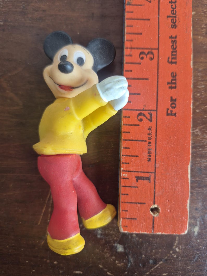 K&ouml;nnte beinhalten: Eine Vintage-Mickey-Mouse-Figur mit gelber Jacke, roter Hose und gelben Schuhen. Die Figur hat schwarze Ohren und wei&szlig;e Handschuhe. Ein Holzlineal, in Zoll markiert, befindet sich neben der Figur. Auf dem Lineal steht der Text "For the finest select".
